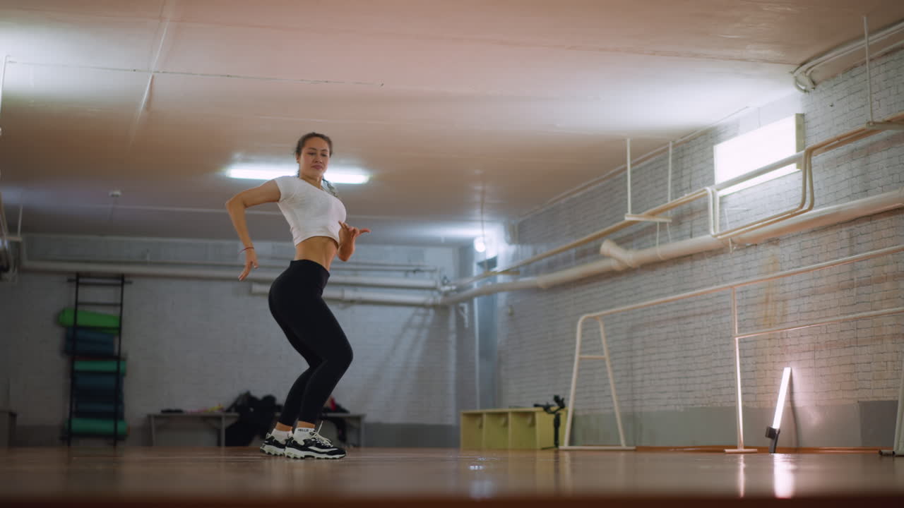 Woman in white top and black leggings dancing inside fitness studio, moving body with energy and rhythm, showing confident graceful performance, focus, and passion on polished wooden floor