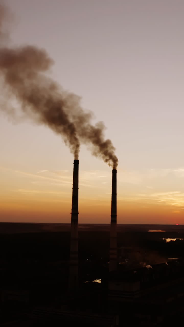 Manufacturing near the river at darkness. Two pipes with dirty smoke going into the air on the nature background in the evening. Vertical video