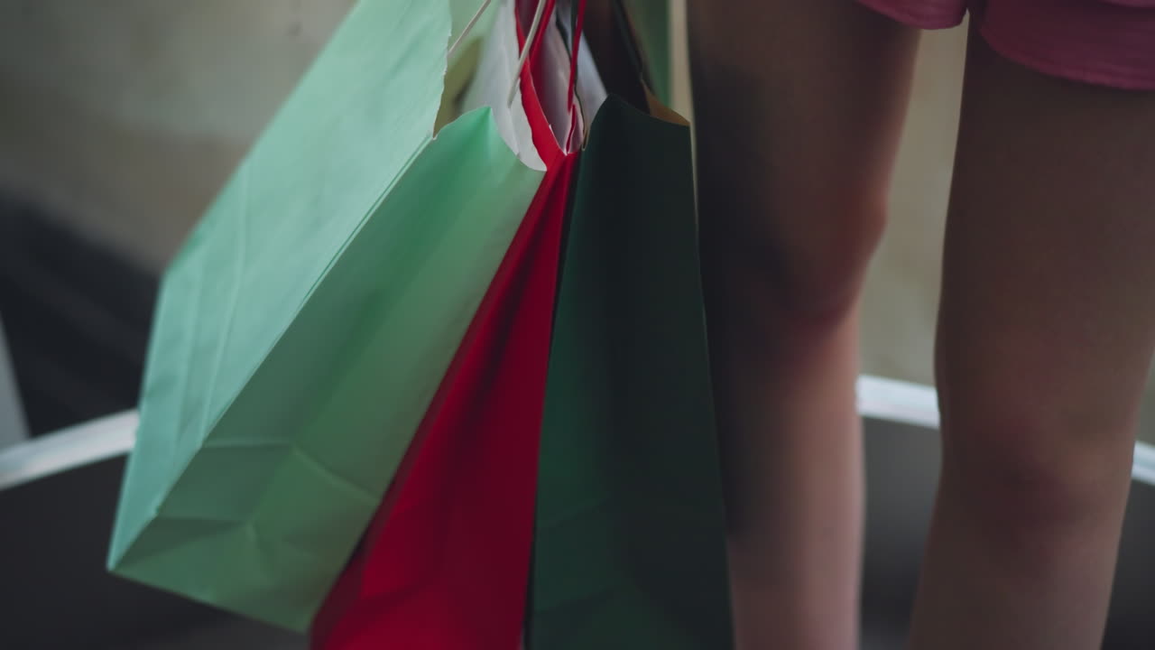 vista trasera de una dama con un vestido rosa de pie dentro de un ascensor, sosteniendo tres bolsas de compras en su mano, las bolsas de compra incluyen tonos de rojo, verde y verde menta, con iluminación suave