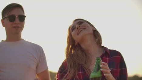 una pareja amorosa está disfrutando de una fiesta al aire libre de verano con amigos. una joven está bailando con cerveza en camisa a cuadros roja con su novio en camiseta blanca y gafas de sol negras.