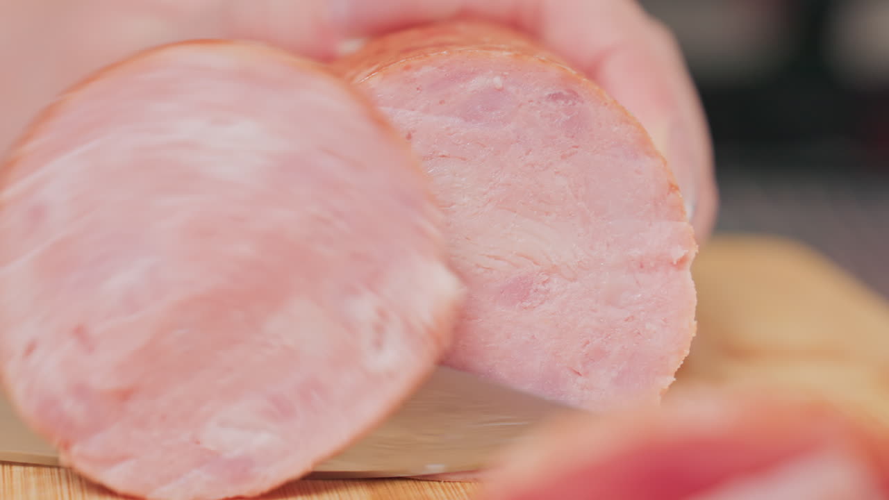 extreme close up person slicing round pink meat sausage with sharp kitchen knife on wooden cutting board, focusing on texture and detail of meat with soft background blur