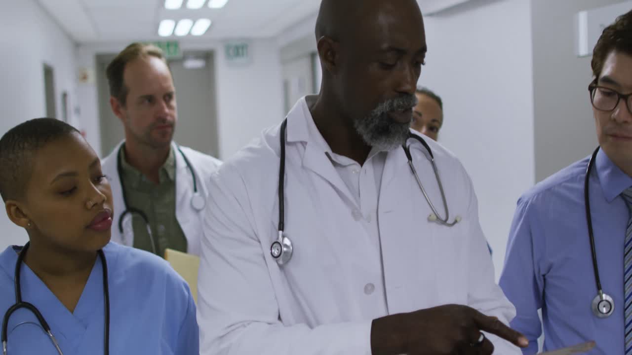 Diverse group of male and female doctors walking in hospital corridor using tablet and talking