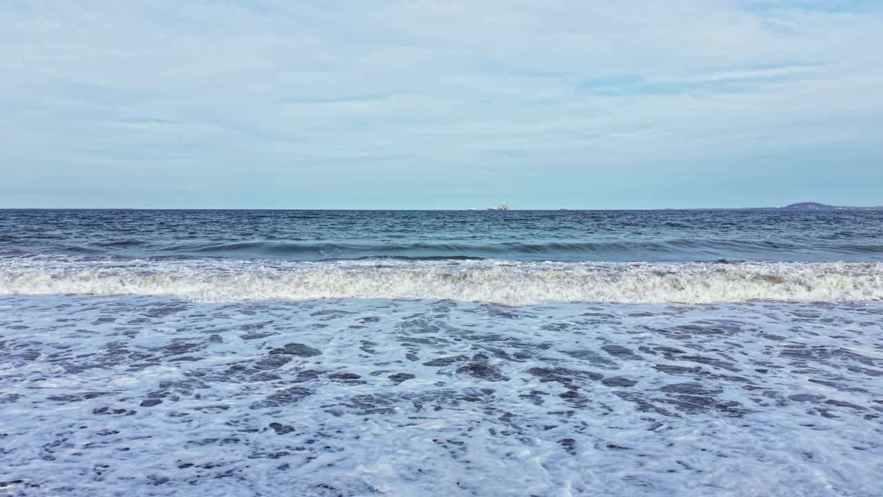 Le onde si infrangono sulla riva mentre la marea sale in una giornata nuvolosa sull'oceano, vicino a una nave lontana e a una terra distante all'orizzonte.