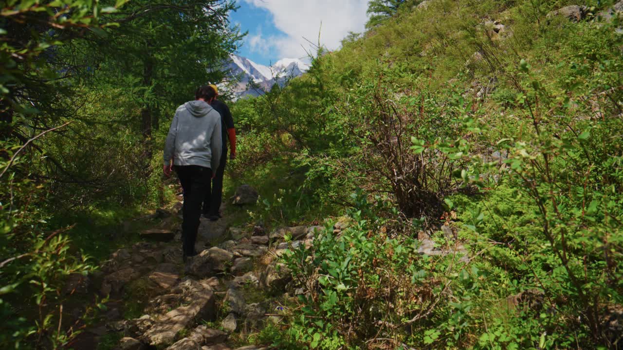 Una persona cammina in salita attraverso una lussureggiante foresta verde. Le montagne si ergono in lontananza sotto un cielo blu. Il sentiero si snoda tra fitti arbusti e rocce. L'escursionista avanza con passo fermo, concentrato sul percorso davanti a sé.