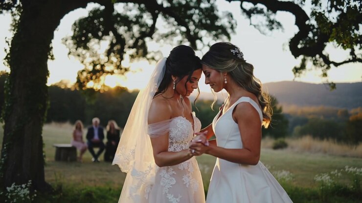 Handholding brides moving closer under oak at sunset, sharing vows in wedding gowns and lace veil