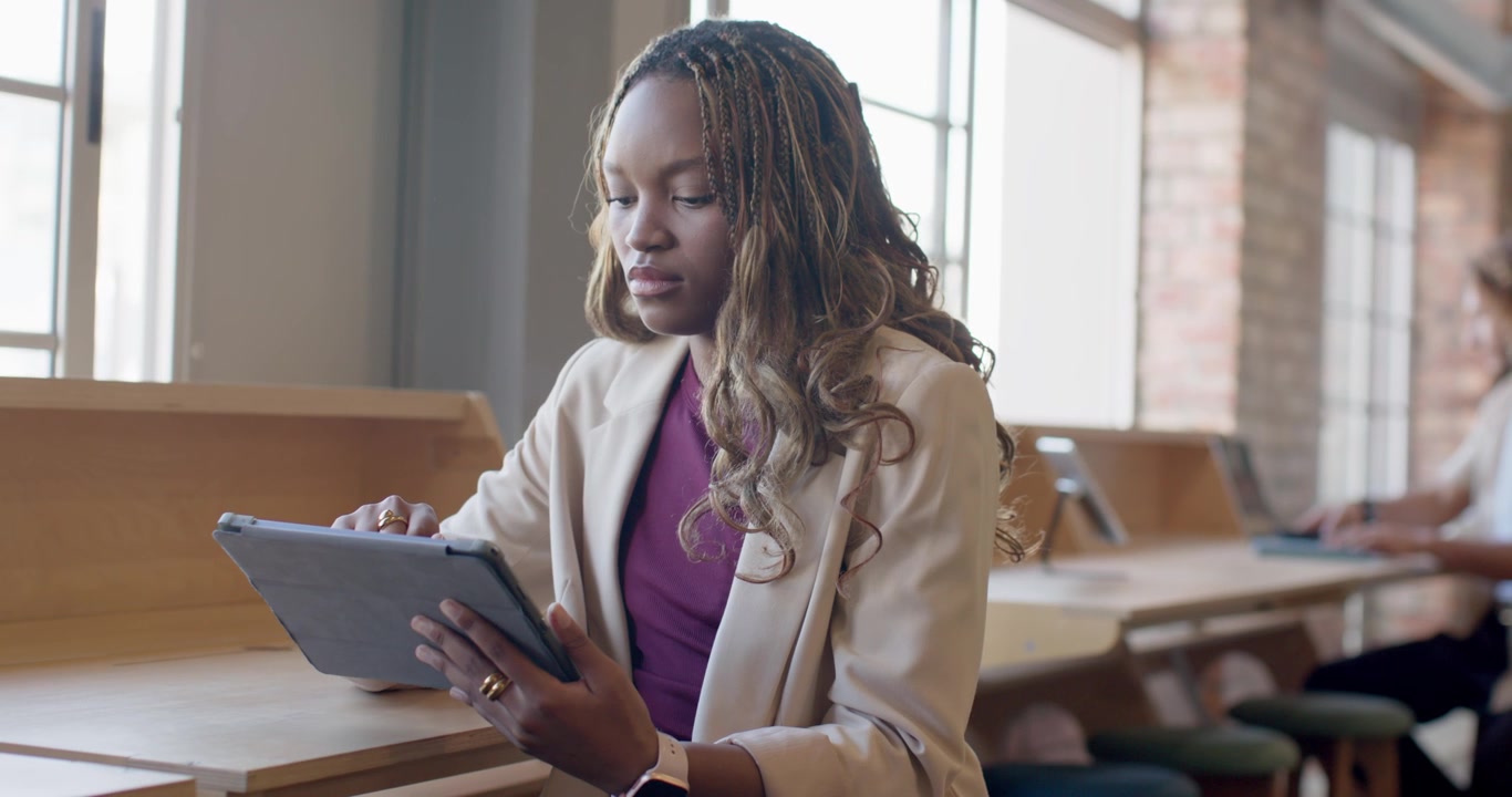 African woman wearing beige blazer at counter by windows holding tablet, tapping, reviewing work
