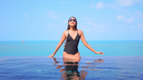 Mujer-Joven-En-Traje-De-Baño-Negro-Sentada-En-El-Borde-De-La-Piscina-Infinita-Y-Tocando-El-Marco-De-Las-Gafas-De-Sol-En-El-Fondo-Del-Paisaje-Marino-Turquesa-En-Un-Día-Soleado-Vista-Frontal-En-Cámara-Lenta