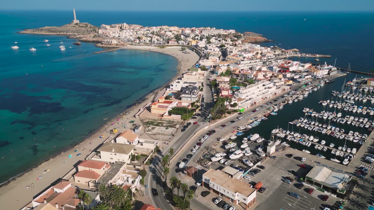 A dolly reverse aerial view of Sea port Marina with a litghhouse in the cliff on the coastal of Cabo de Palos, Murcia region - a touristic beach summer vacation in Spain