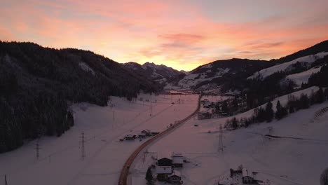 Aerial-shot-of-a-snow-covered-mountain-town,-winding-road-through-the-valley-during-sunset