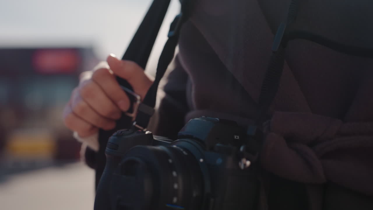 Closeup street photographer holding camera strap and lens, wearing dark coat with tied belt, camera hanging at hip with lens cap attached, blurred urban bus background, soft bokeh and muted light