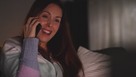 close up of smiling woman sitting on sofa at home at night talking on mobile phone 1