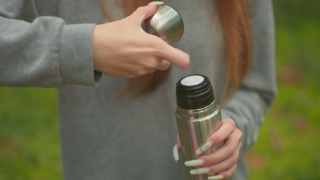 close-up of young woman in grey sweater and long hair cascading down, pressing inner cover of thermos, pours water into lid, highlighting delicate, long nails in a forest setting