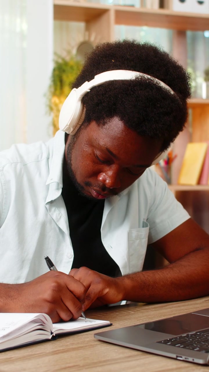 African american young man on home table studying with headphones laptop and notes for exams indoors