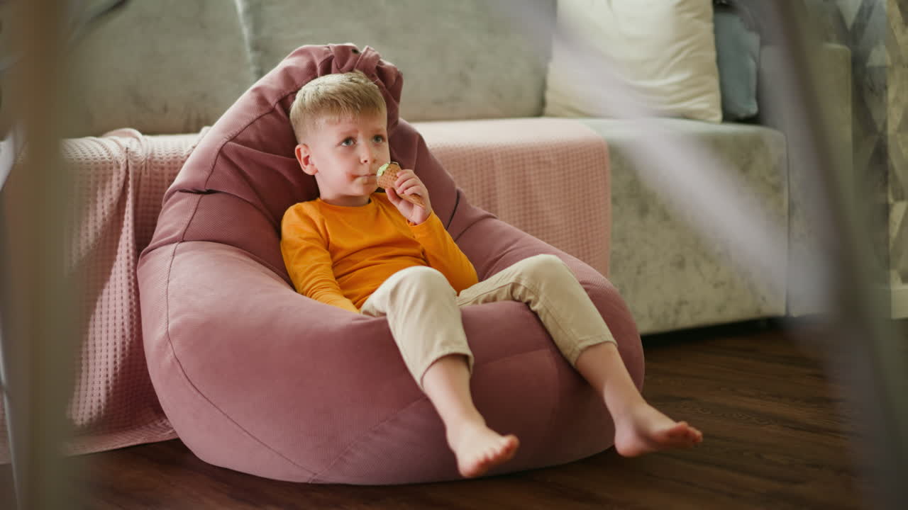 Cute young boy lounges comfortably on large pink beanbag indoors, joyfully licking mint ice cream cone while stretching legs playfully, with blurred metal stand structure partially visible
