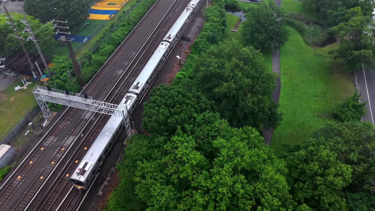 An aerial view of a Long Island Rail Road train approaching the Valley Stream station on a cloudy day. The drone camera is tilted downward and dolly in following the slow moving train.