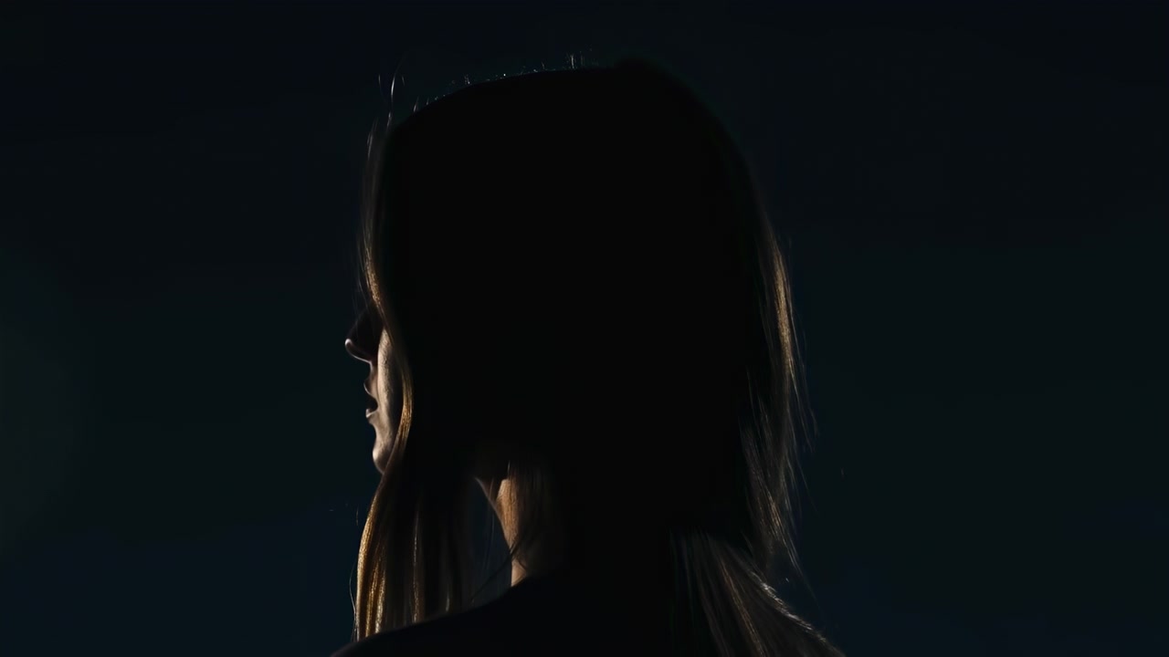Shadowed side profile stays framed against a deep blue-black background as narrow warm backlight outlines long hair, nose, lips, and neck during gentle changes in head position.