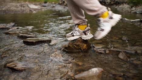 shooting close up: a girl in white sneakers crosses a mountain river along a special path made of stones, her boyfriend who is standing on the other side of the river helps her