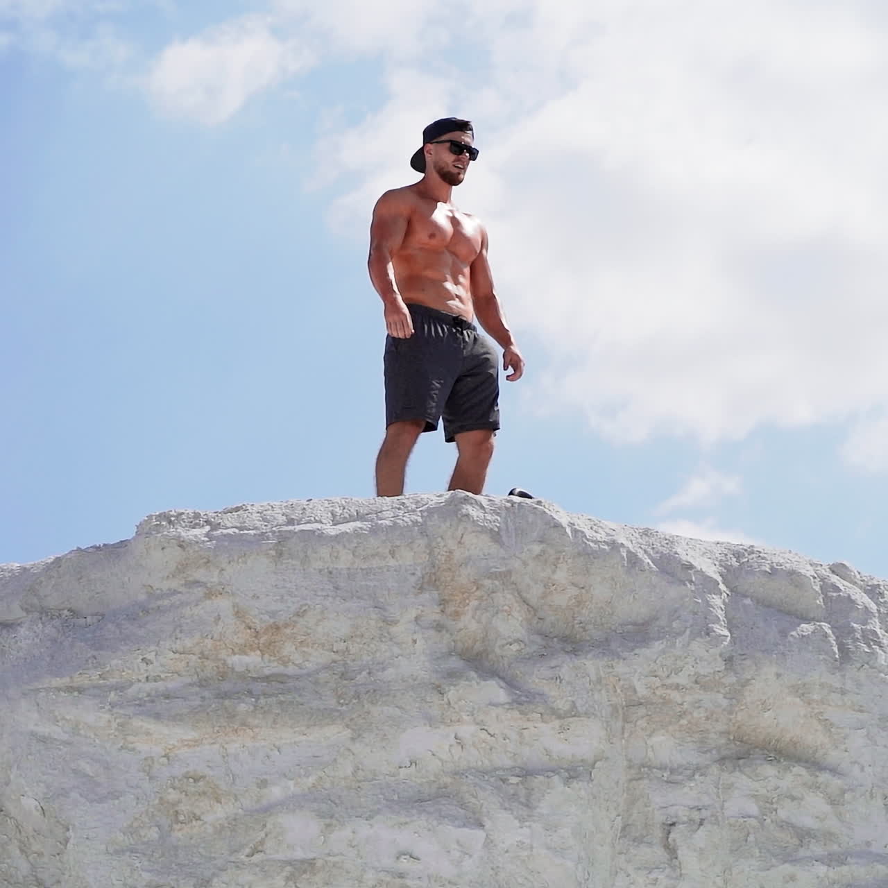 Shirtless bodybuilder with sunglasses and cap on the hill in a sunny day. Strong athlete with muscular body under blue sky outdoors.