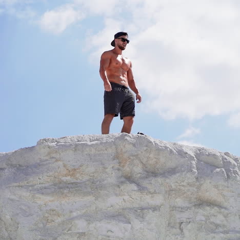 shirtless bodybuilder with sunglasses and cap on the hill in a sunny day. strong athlete with muscular body under blue sky outdoors.