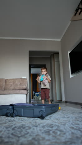 cute caucasian toddler holding a bunch of his clothes. kid tosses his things up cheering happily. packing a suitcase at home. low angle view. vertical video.