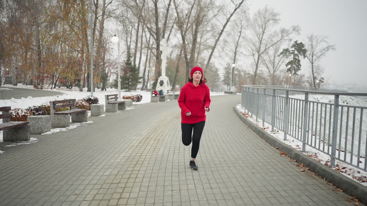 Freelancer jogging along snowy park pathway with bag placed on distant bench, surrounded by serene winter scenery, trees, and bushes, near iron railing and lamp posts showcasing fitness