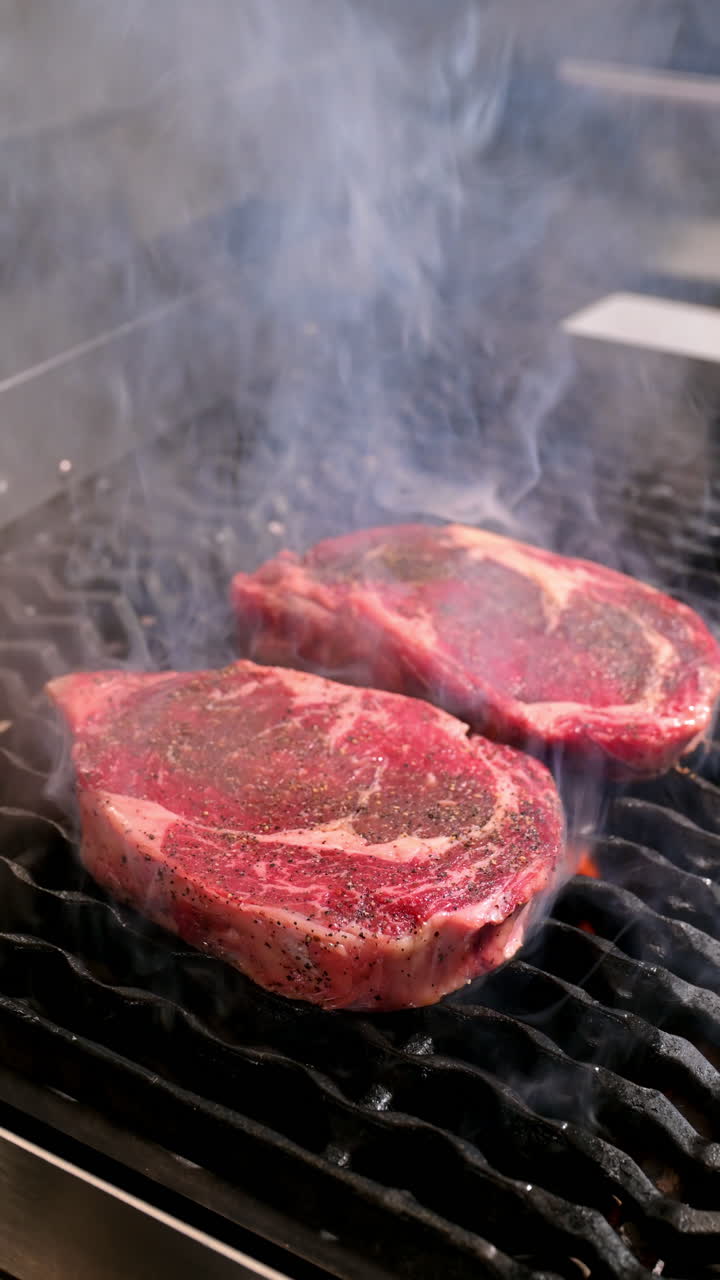 Two raw steaks grilling on a smoky barbecue. Close-up of two raw beef steaks on a smoky hot grill with seasoning and flame