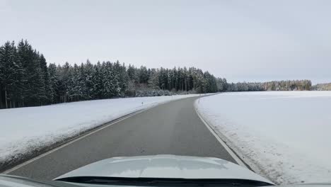 Drivers-front-view---driving-a-car-through-a-snowy-landscape-next-to-a-forest-with-snow-covered-trees-along-white-powder-fields,-beautiful-winter-scenery-from-the-windshields-view