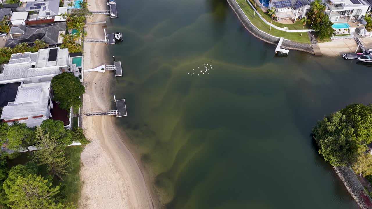 Drone footage glides above a riverfront neighborhood with luxury houses, private docks, sandy shoreline, and lush greenery under bright daylight