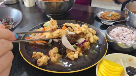 close-up of a hand taking with chopsticks crispy sweet and sour pork tangsuyuk, served with multigrain rice and assorted korean side dishes on a restaurant table