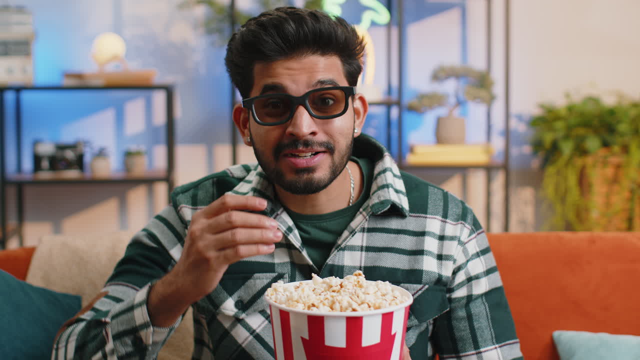 hombre indio se sienta en el sofá comiendo palomitas de maíz y viendo interesante serie de televisión juego deportivo en línea en casa