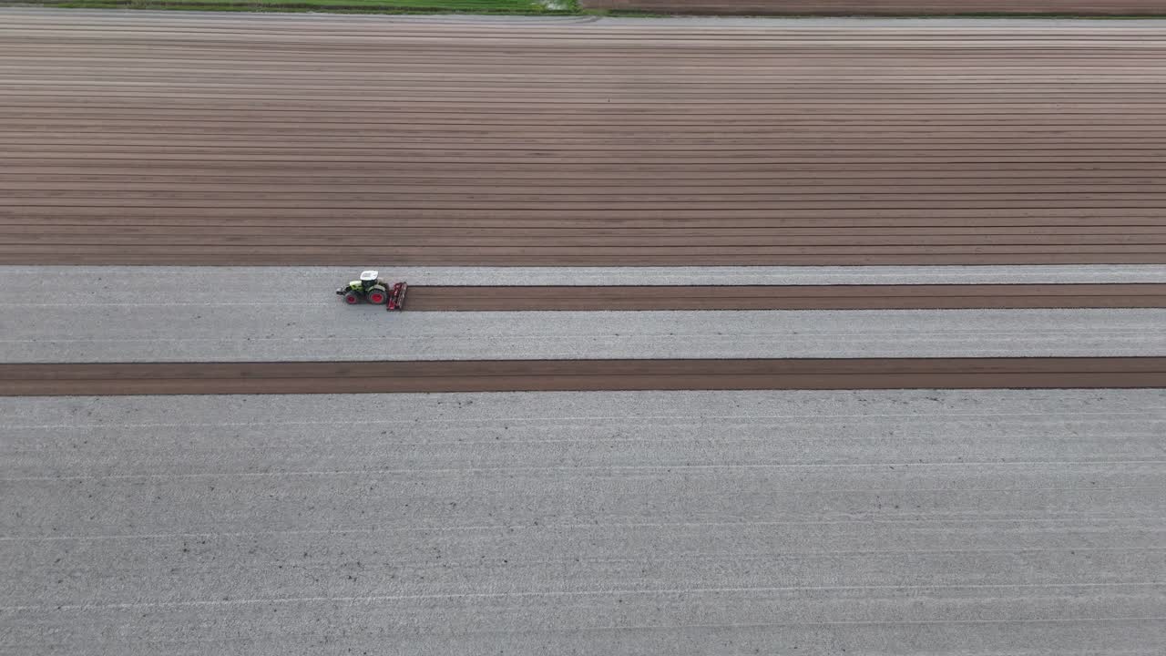 Tractor leveling soil in slow motion, carving uniform dark seed rows beside light-bleached furrows across an expansive field under warm dusk light, drone tracking shot