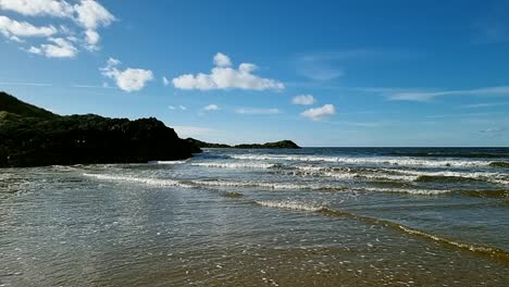 Sunlit-slow-motion-ocean-waves-moving-towards-lush-green-paradise-North-Wales-island