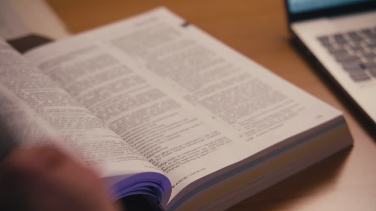 Close-up handheld shot of a man's hand turning pages of a large German reference book at a desk. Regulations text visible, calculator in background