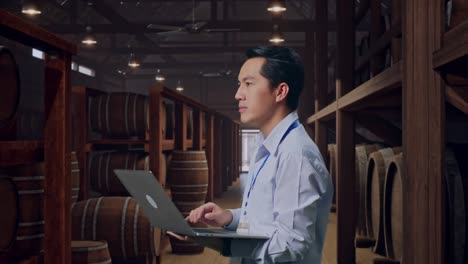 side view of an asian male professional worker standing in cellar of winery, observes by looking up before he come to concentrating on the laptop and keep on typing