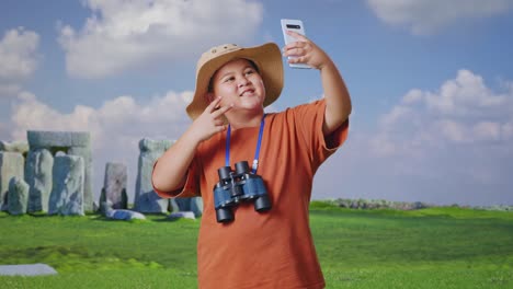 asian boy with a hat and binoculars taking a selfie on smartphone while traveling in stonehenge. boy researcher examines something, travel tourism adventure concept