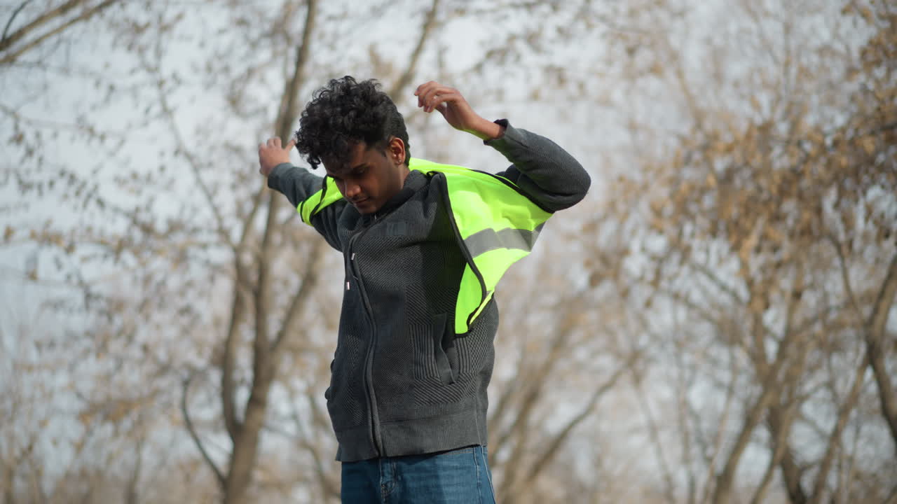 Man in outdoor setting putting on bright neon green reflective safety vest, preparing for environmental work or volunteer cleanup in park during early spring with bare trees and muted background scenery