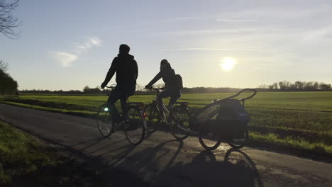 Toma-En-Cámara-Lenta-Que-Muestra-La-Silueta-De-La-Familia-En-Bicicleta-Con-Un-Transportador-De-Niños-Montando-En-Un-Camino-Rural-Durante-La-Puesta-De-Sol