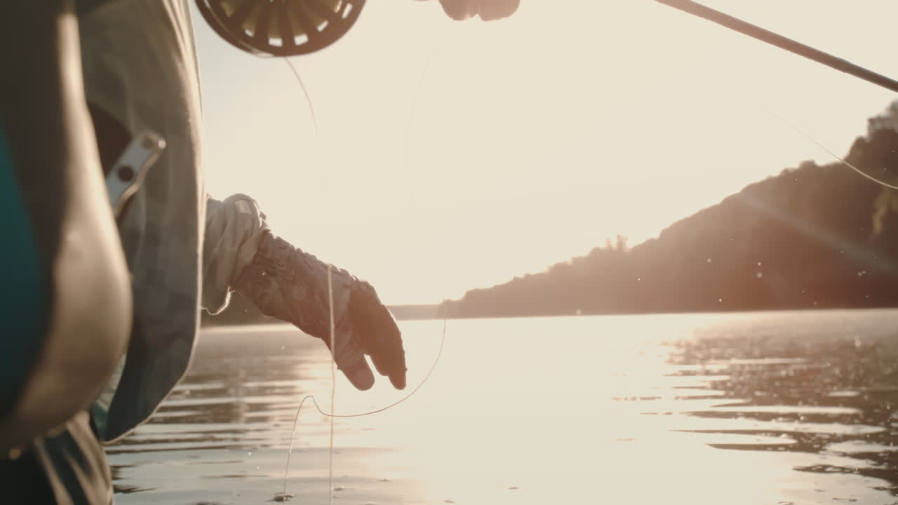 Slow motion of fly fisherman pulling line, revealing gear pack on riverbank as golden hour sunset reflects on rippling water during fly fishing action