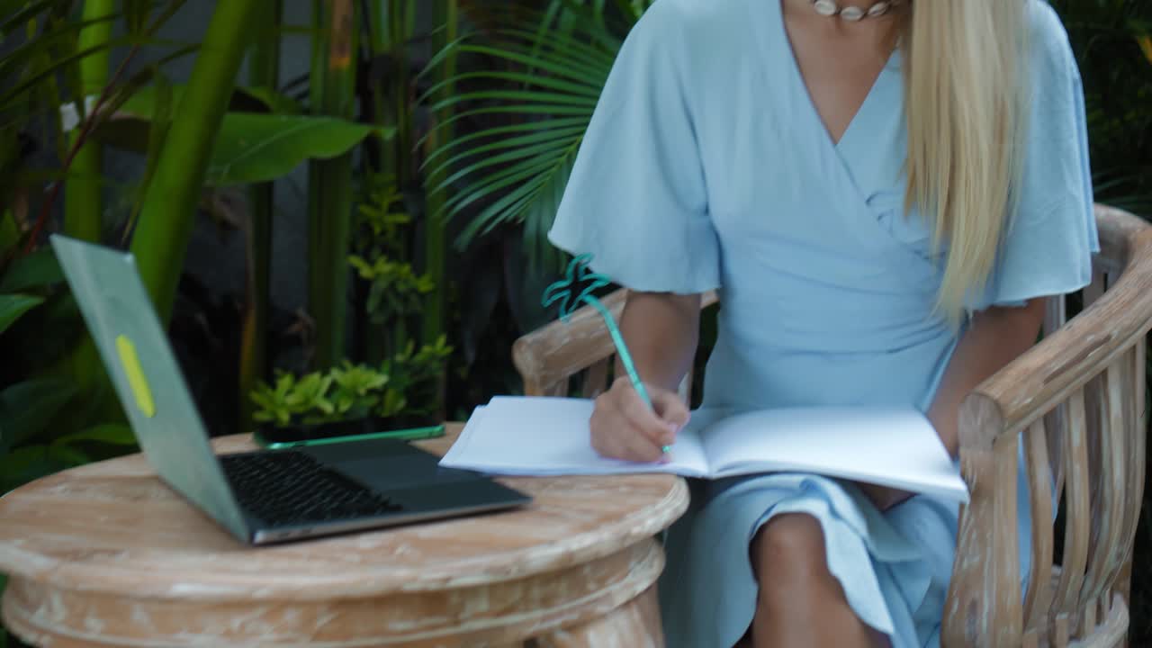 A young European girl in a blue dress remotely online working on laptop and looking into the screen on the backyard with green plants