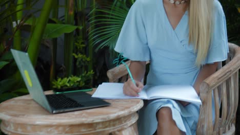 una joven europea en un vestido azul remotamente en línea trabajando en una computadora portátil y mirando a la pantalla en el patio trasero con plantas verdes