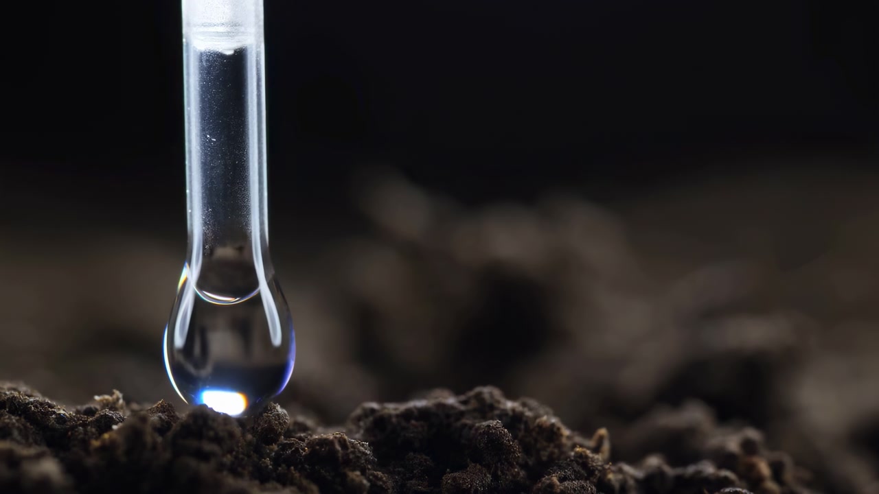 Goccia trasparente scende lentamente dalla punta di una pipetta e tocca la superficie irregolare del terreno, illuminata da una luce focalizzata mentre lo sfondo poco profondo rimane scuro e leggermente sfocato.