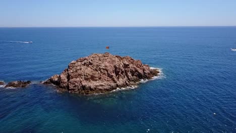 Flying-around-an-island-with-a-flag-on-the-Mediterranean-sea-with-a-boat-behind-it