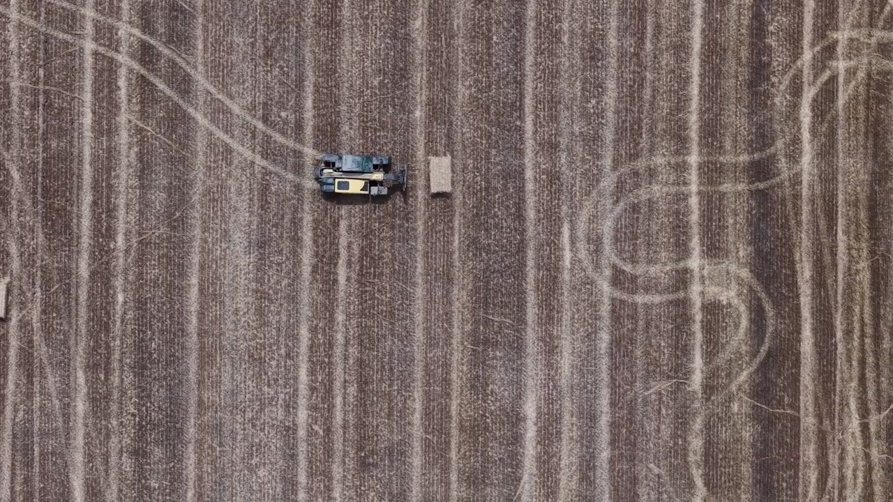Un trattore agricolo giallo lavora un vasto campo arato da una prospettiva aerea, con i segni dei pneumatici che formano motivi sorprendenti sul terreno asciutto