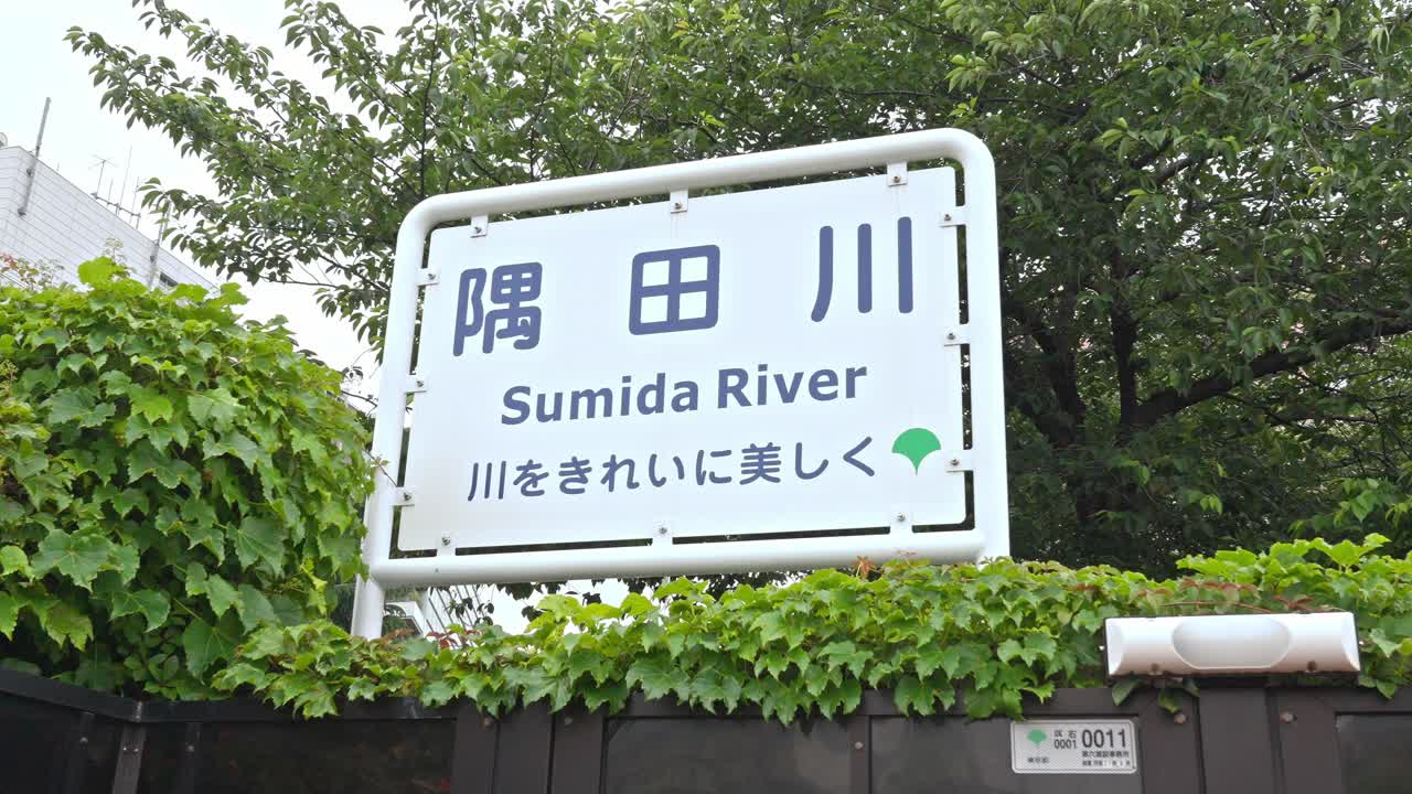 A "Sumida River" signboard with the message Keep the river clean and beautiful surrounded by lush green foliage.