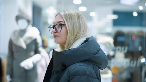 russian lady with glasses passing by well-lit clothing store in mall, clothes displayed on mannequins visible through glass with soft bokeh light effect, creating an artistic atmosphere