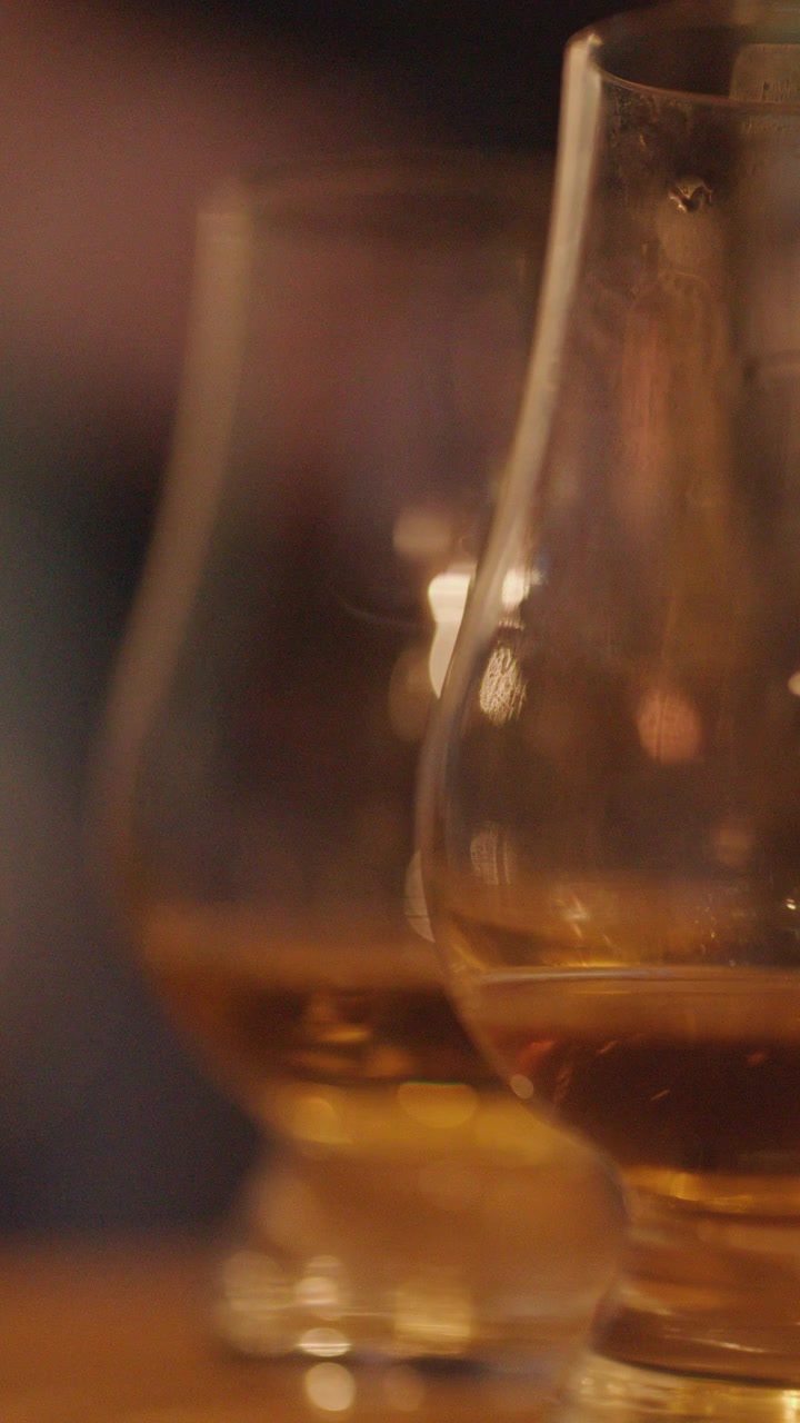 Due bicchieri da whisky con liquido ambrato poggiano su una superficie di legno, catturati con un'illuminazione morbida e calda, con una profondità di campo ridotta e un movimento minimo della fotocamera.