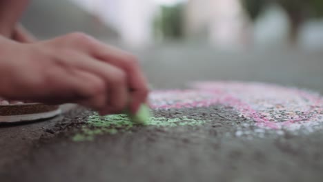 Handheld-Close-Up-side-shot-of-a-girl-child-sitting-on-the-sidewalk-drawing-a-mushroom-on-the-street-asphalt-pavement-with-green-colourful-chalk-in-glowing-evening-light