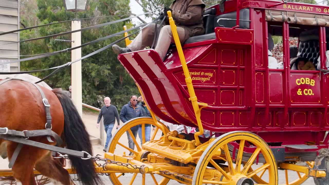 A vibrant red Cobb & Co. carriage pulled by Clydesdale horses passes by spectators on a tree-lined street.
