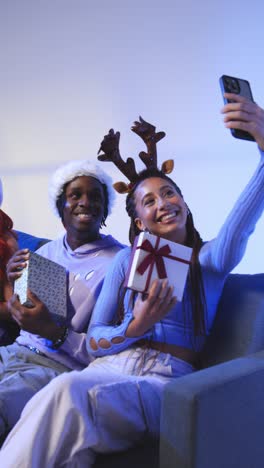 Vertical-Video-Studio-Shot-Of-Gen-Z-Friends-At-Christmas-Sitting-On-Sofa-Wearing-Santa-Hat-And-Reindeer-Antlers-Taking-Selfie-On-Mobile-Phone