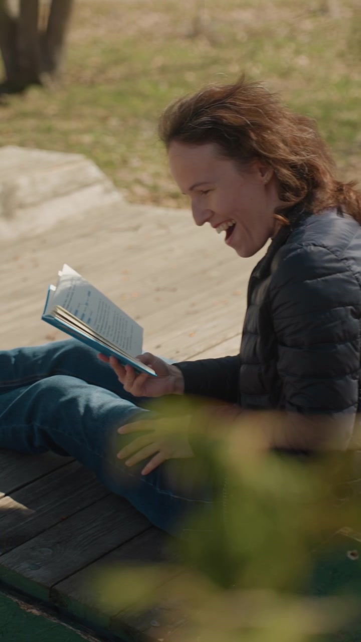 Primo piano di una donna che legge un libro su una panchina di legno in un parco con sfondo di fogliame verde ed erba. Il libro è aperto, tenuto dalla mano, mostrando le pagine di testo. Una luce naturale soffusa illumina la scena creando un'atmosfera rilassante.